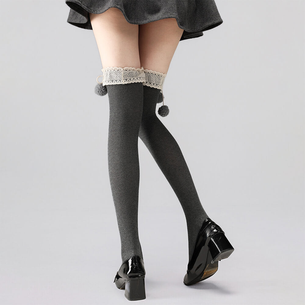 Gray knee-high socks with lace trim and pom-poms worn with black shoes on a light gray background