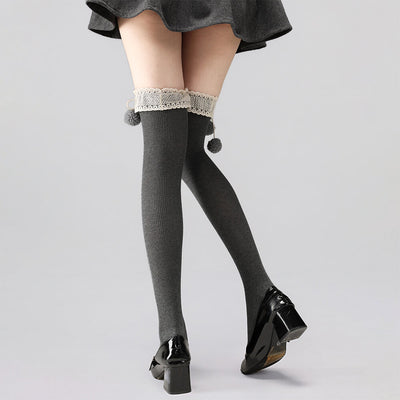 Gray knee-high socks with lace trim and pom-poms worn with black shoes on a light gray background