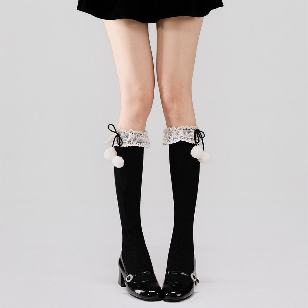 Black calf socks with lace and pom-pom details worn with black shoes on a light gray background