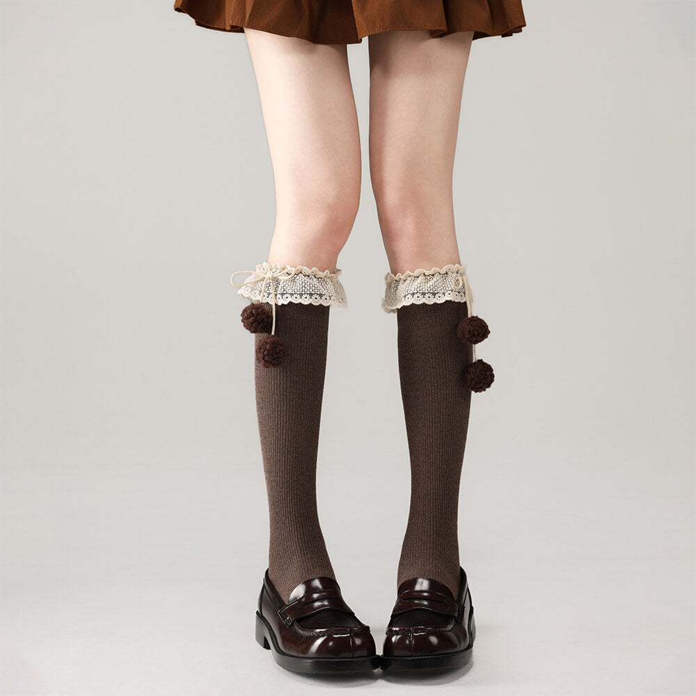 Brown below knee socks with lace and pom-pom details worn with black shoes on a plain background