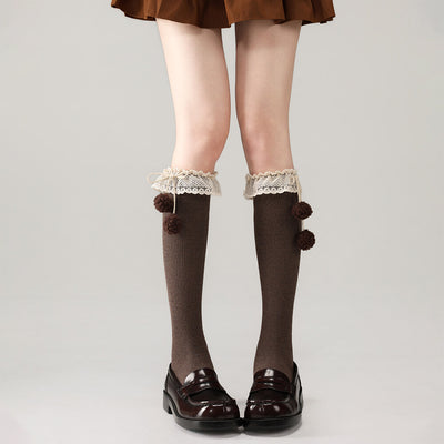 Brown below knee socks with lace and pom-pom details worn with black shoes on a plain background