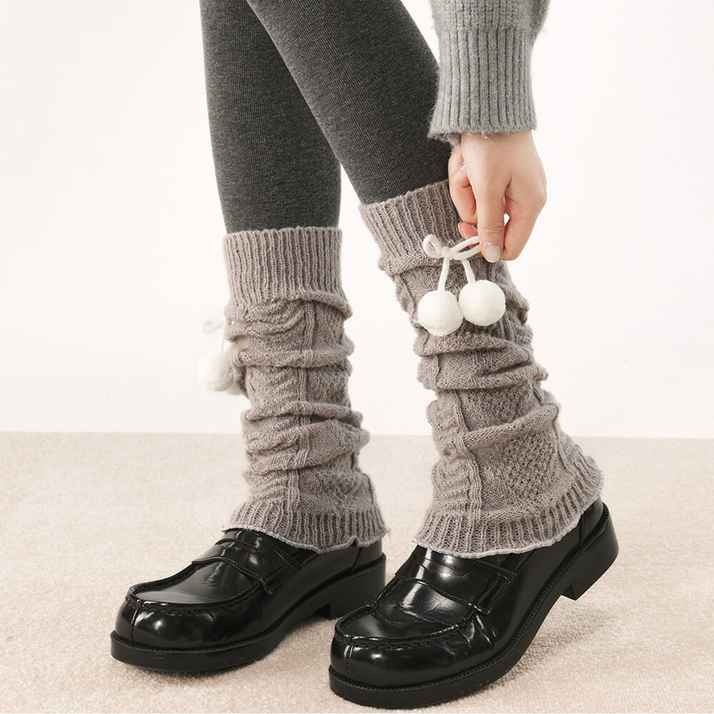 Person wearing gray knitted leg warmers with white pom-poms and black shoes on a light background