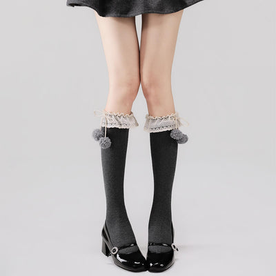 Dark gray calf socks with lace and pom-pom details worn by a person on a light gray background