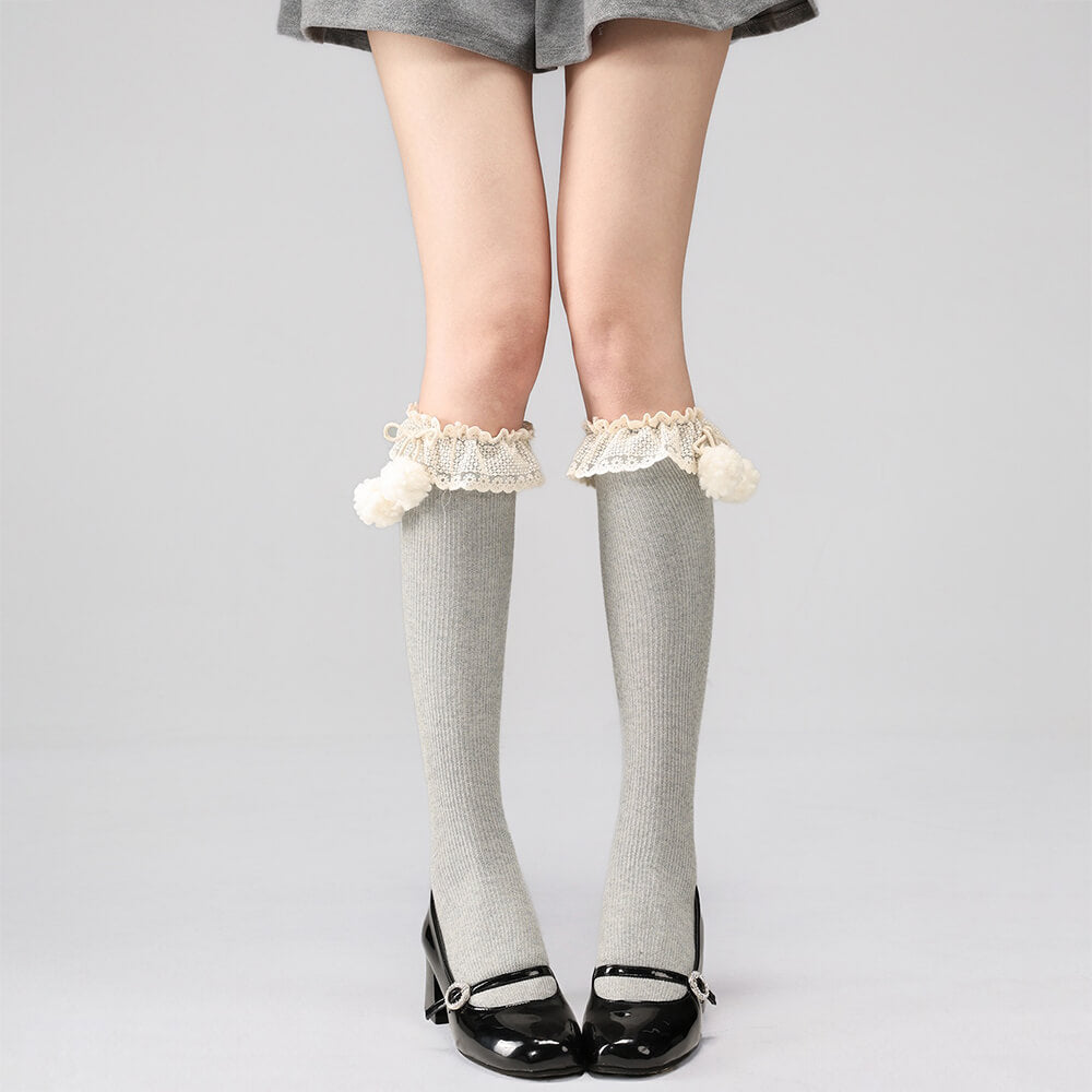 Light gray below knee socks with lace top and pom-poms worn with black shoes on a plain background