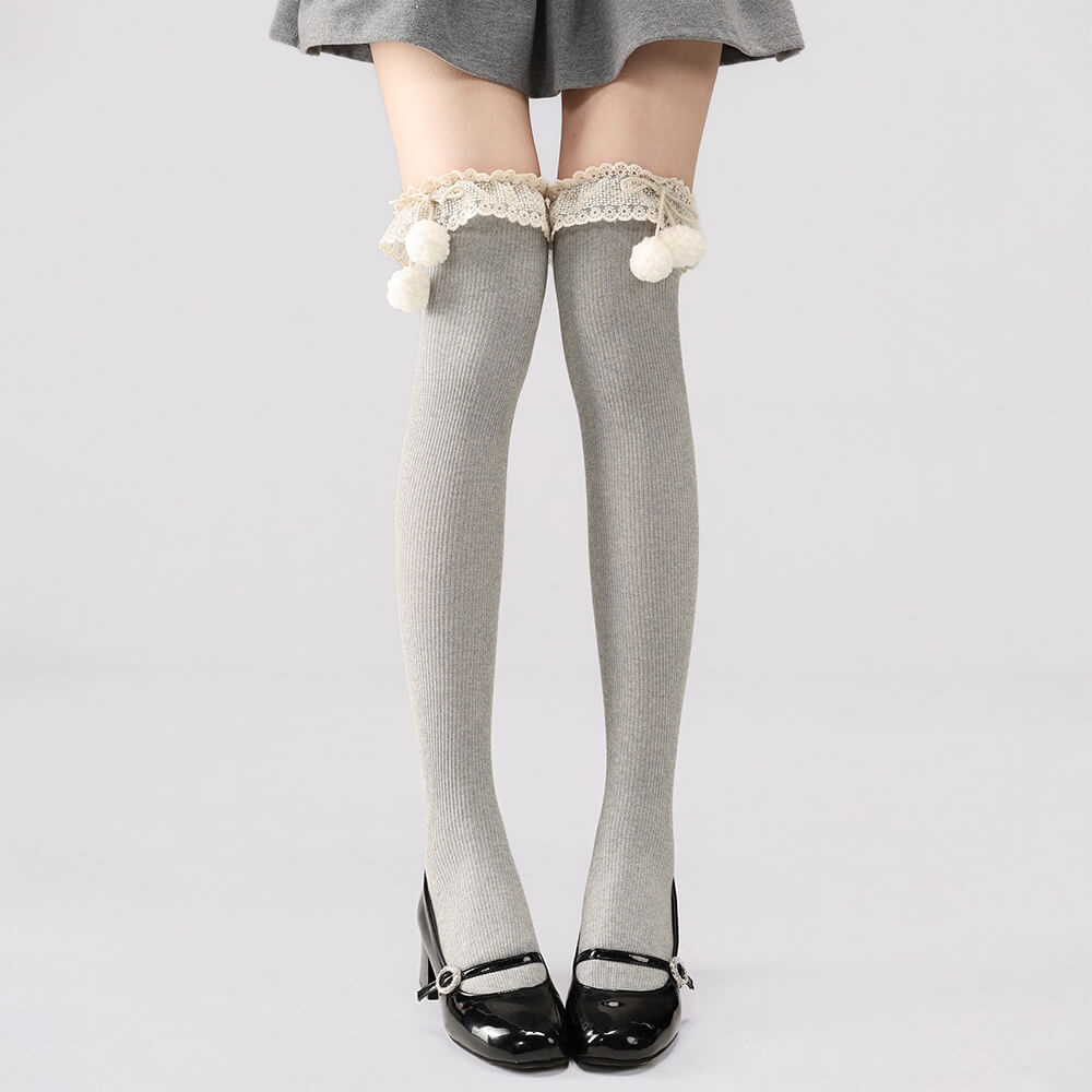 Gray patterned knee-high socks with lace and pom-pom details worn by a person on a light gray background.