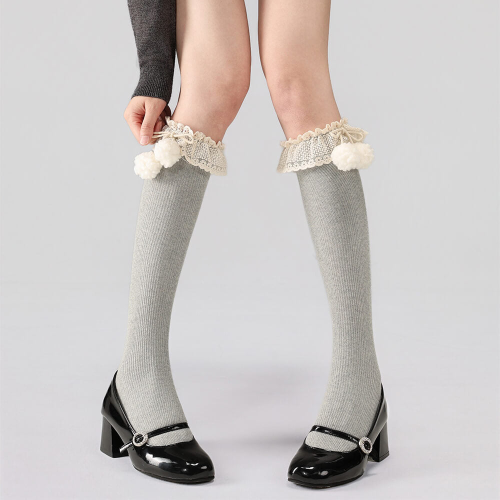 Gray calf socks with lace and pom-pom details worn by a person on a light gray background.