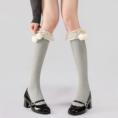 Gray calf socks with lace and pom-pom details worn by a person on a light gray background.