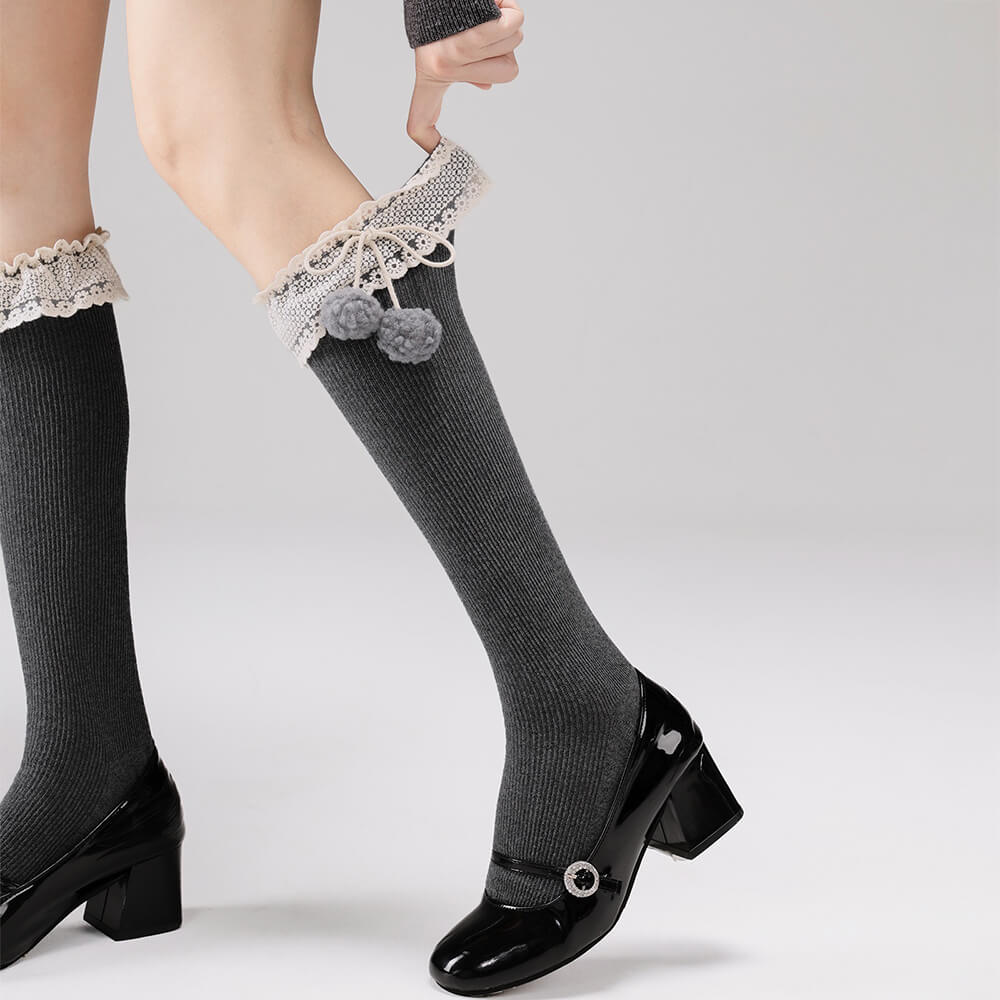Gray knee-high socks with lace top and pom-poms worn with black shoes on a light gray background